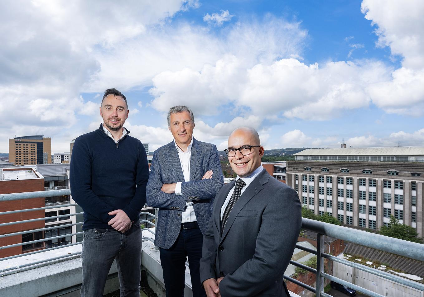 Three men suited standing on balcony overlooking city skyline.