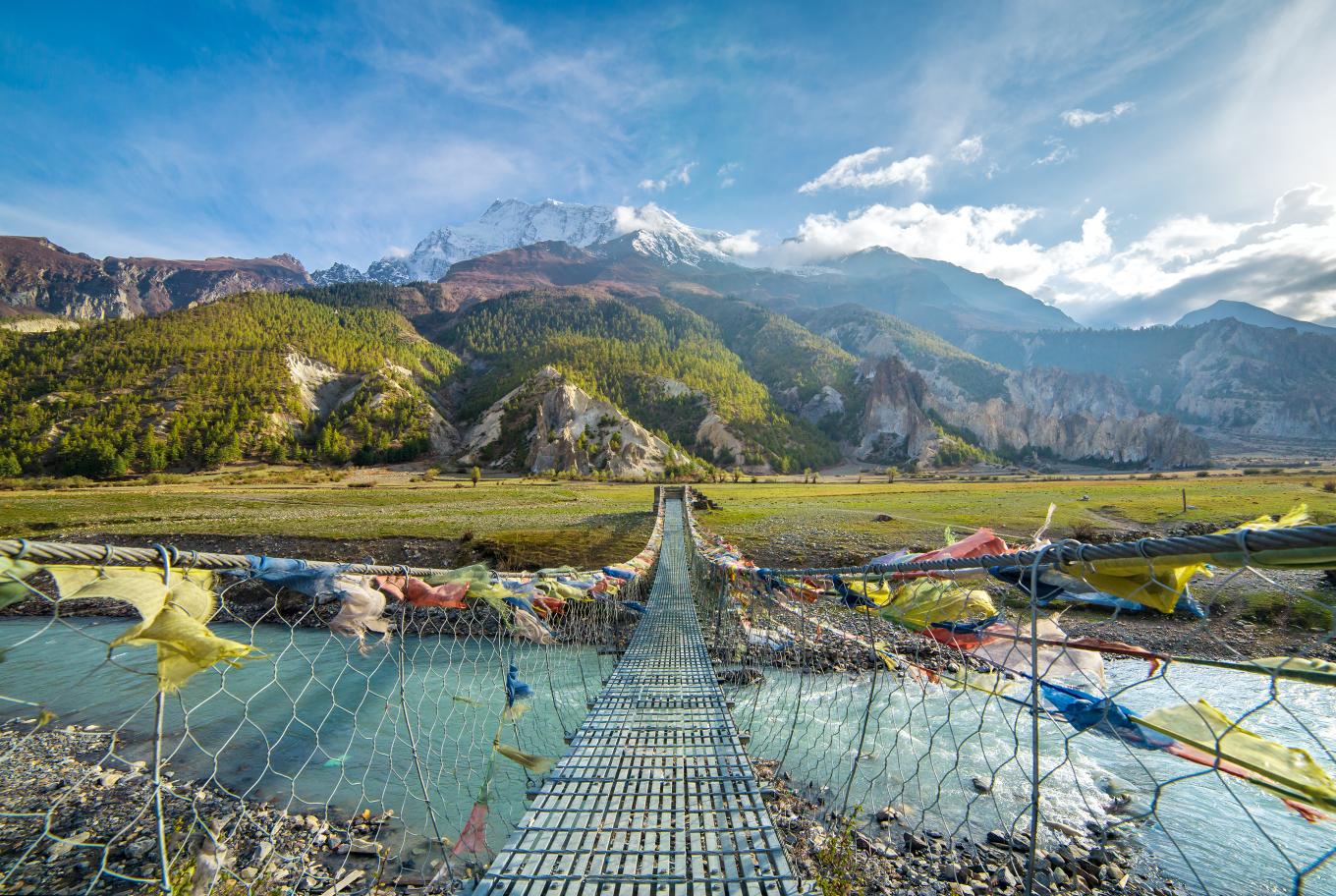 A panaromic views of mountains in the background, a stream in the foreground with a rope bridge crossing over it