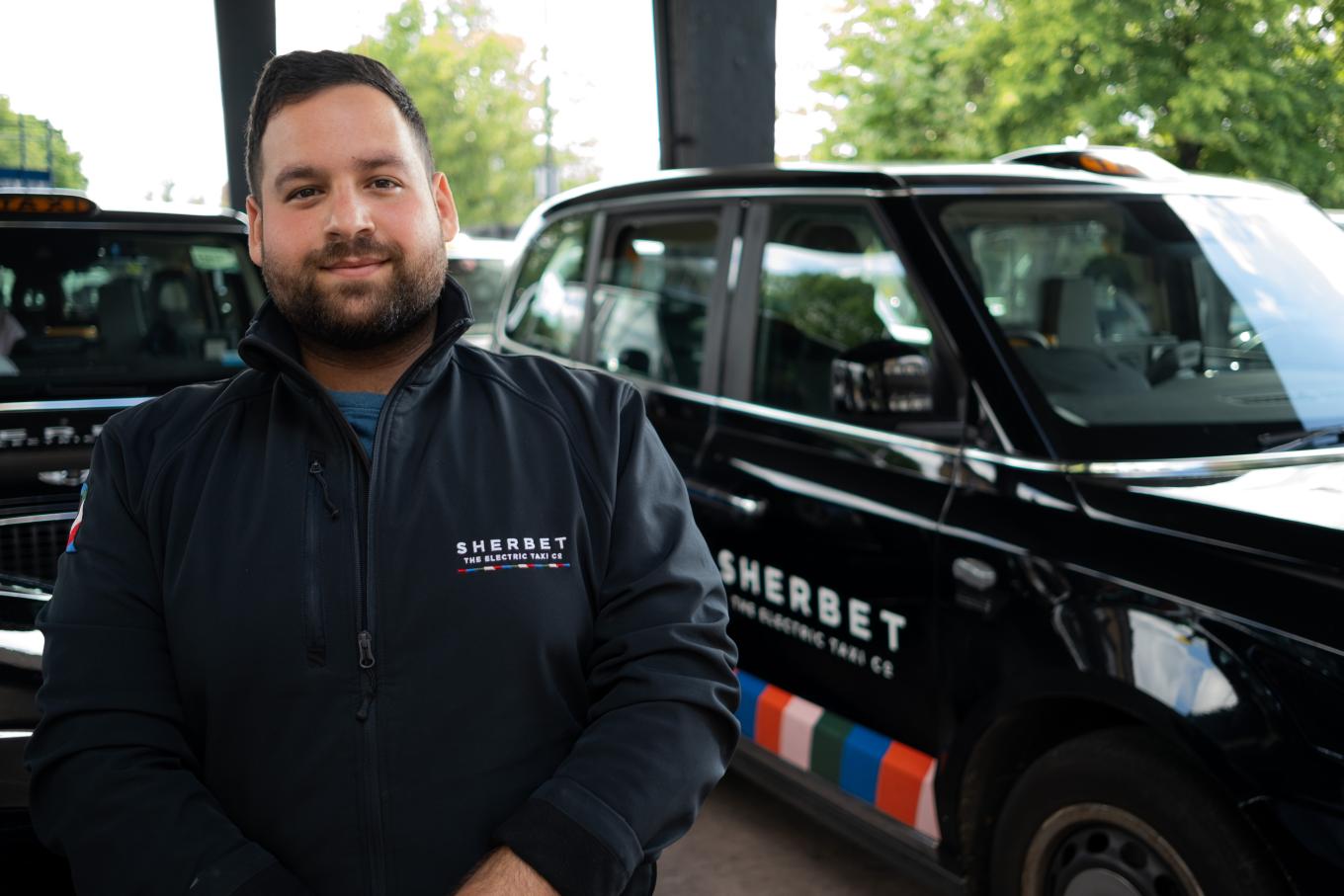 A man stands in front of a black cab with the branding Sherbet Taxis