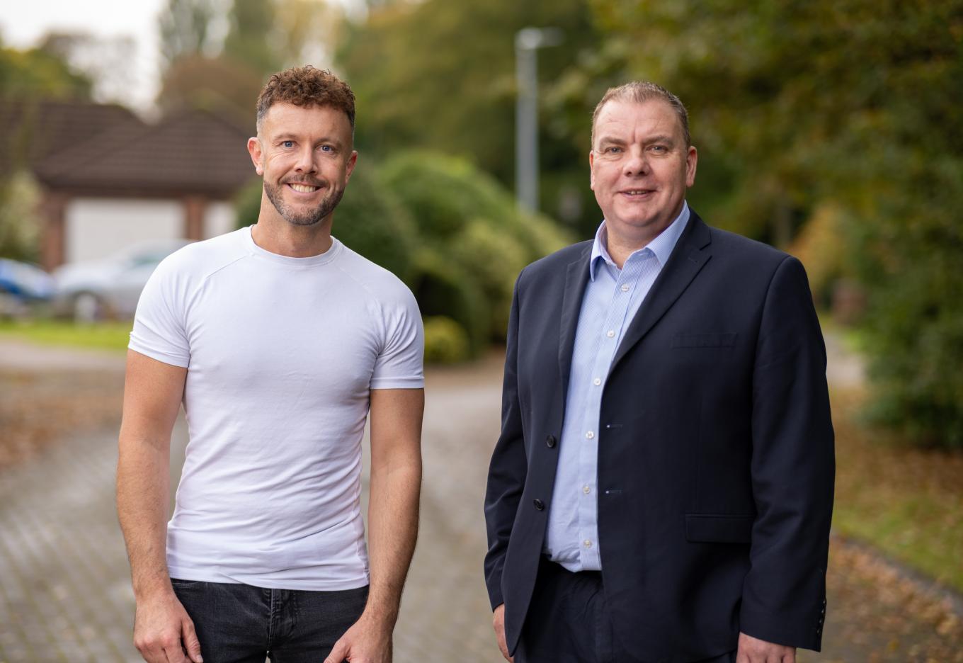 Two men standing on a paved path outdoors. One is in a white t-shirt and the other in a suit, with trees and a house in the background.
