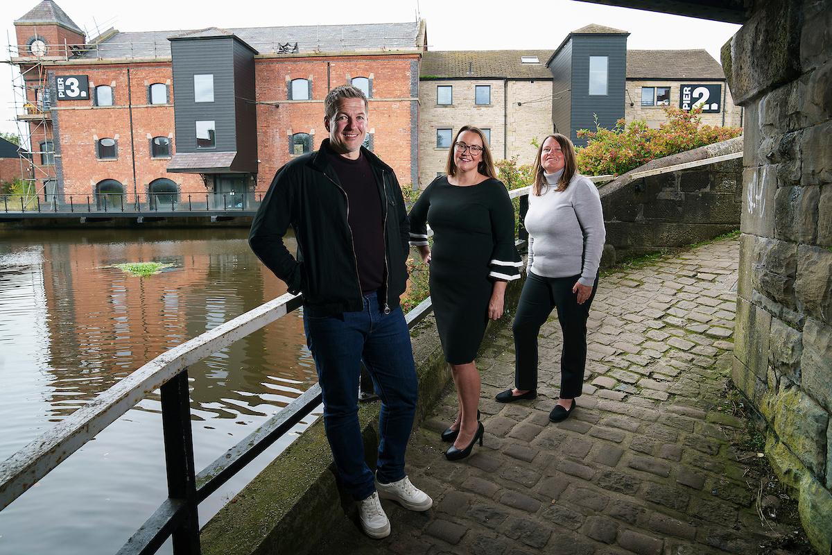 Three people, two women and one man, stand on a cobbled walkway beside a calm canal with buildings in the background