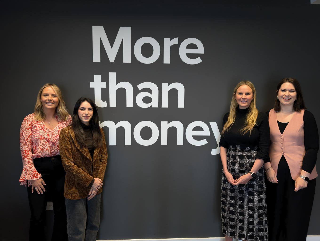 A group of four women stands proudly before a sign stating "More Than Money,"
