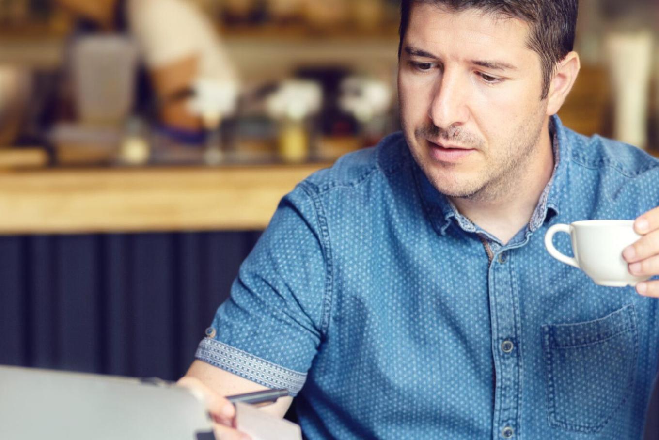 Casually dressed man sat in a coffeeshop drinking coffee and working on a laptop device