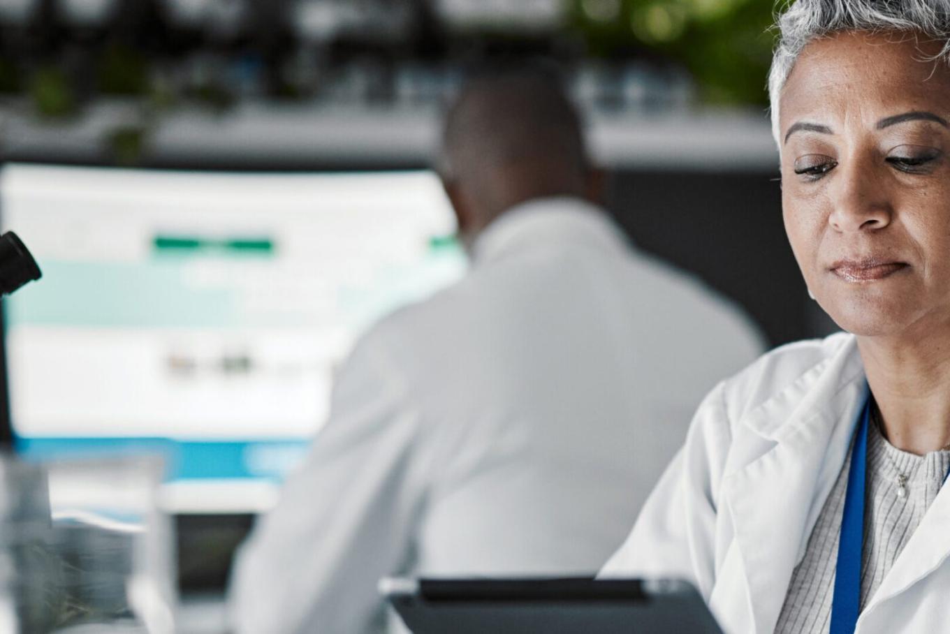 A male and female scientist or laboratory technican in lab conditions with a microscope in sight.